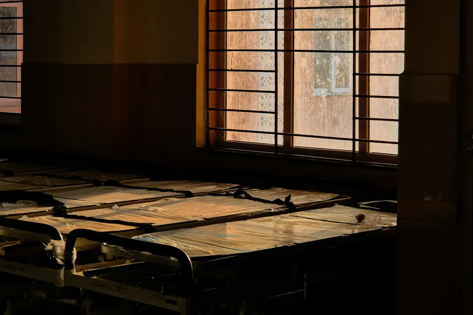 Empty hospital beds in a dim ward lit by a window