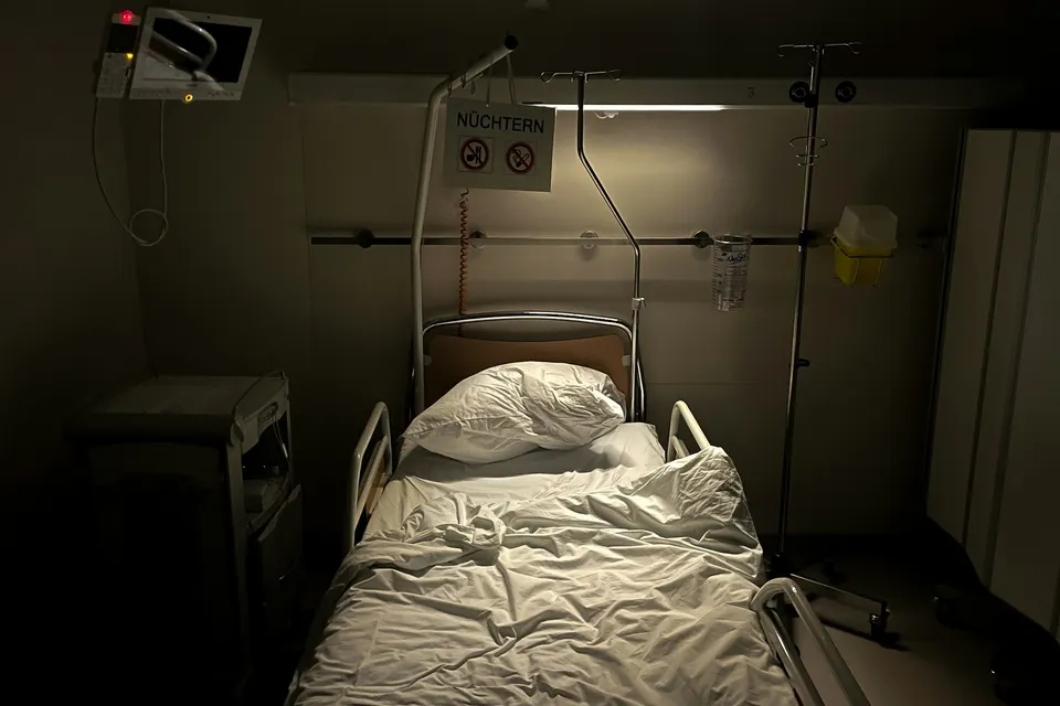 An empty hospital bed in a dim room next to an IV pole and a bedside monitor
